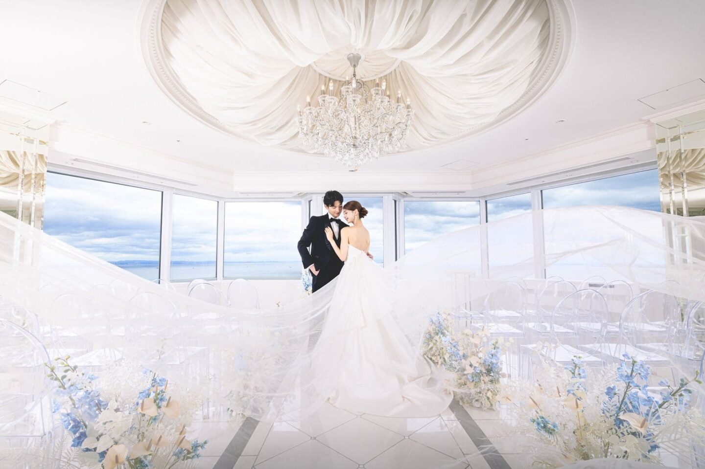 海が一望できる白基調のチャペルで、長いベールをなびかせて寄り添うドレス姿の新婦とタキシード姿の新郎