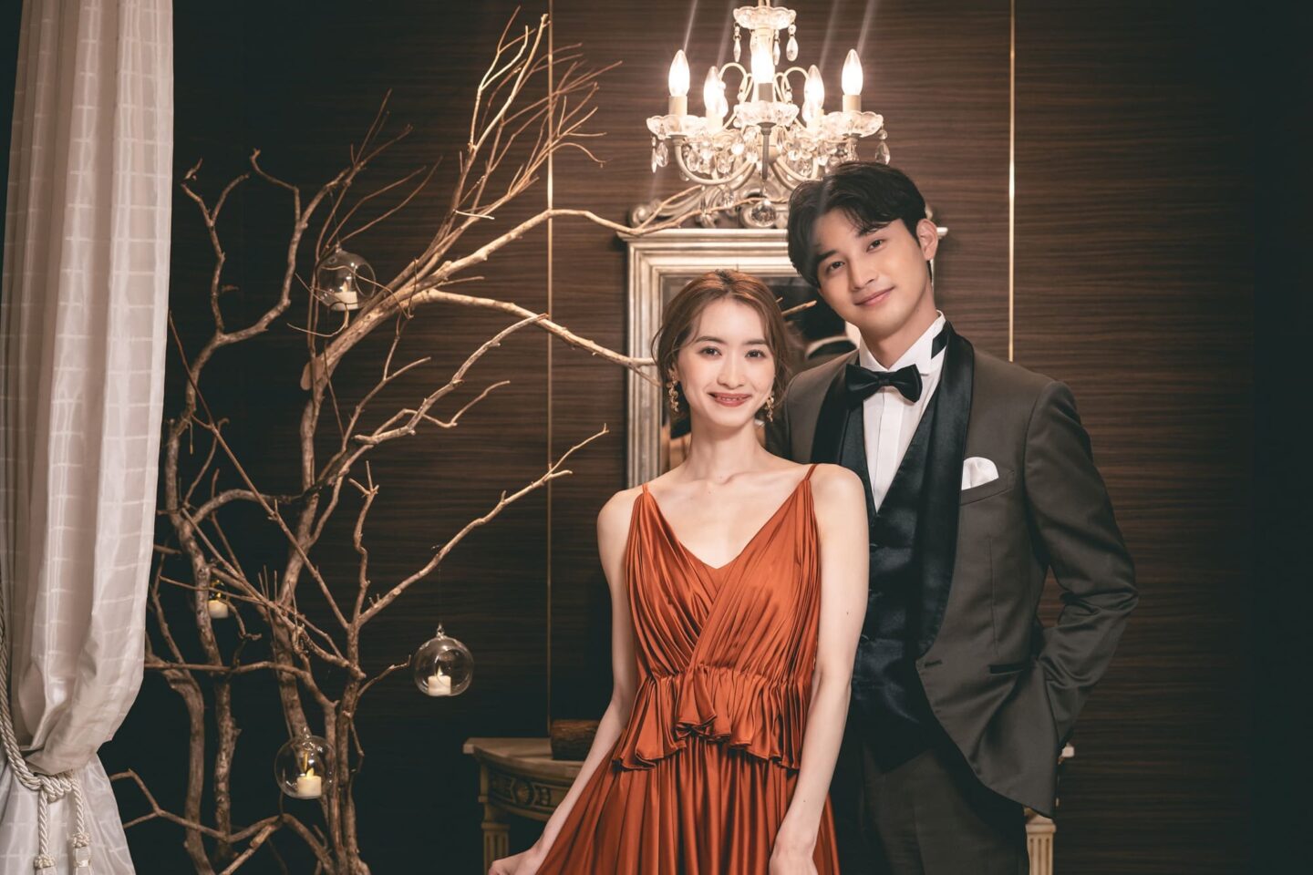 韓国風フォトウェディング|シャンデリアが輝くシックなスタジオで寄り添う、テラコッタドレスの新婦とタキシードの新郎