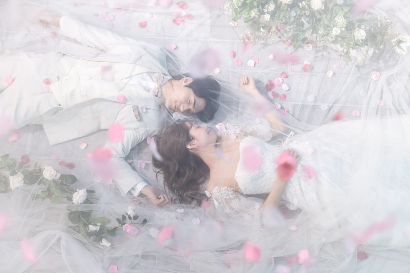 舞い散るピンクの花びらとチュールに包まれ、仰向けに寝そべる白タキシードの新郎とウエディングドレス姿の新婦