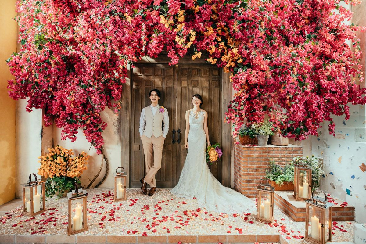鮮やかなピンクの花々に囲まれた木製扉の前に立つ、ウエディングドレス姿の新婦とベージュスーツの新郎