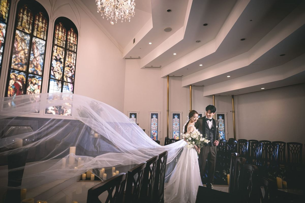 ステンドグラスのあるチャペルでロングベールを広げる新婦と寄り添う新郎